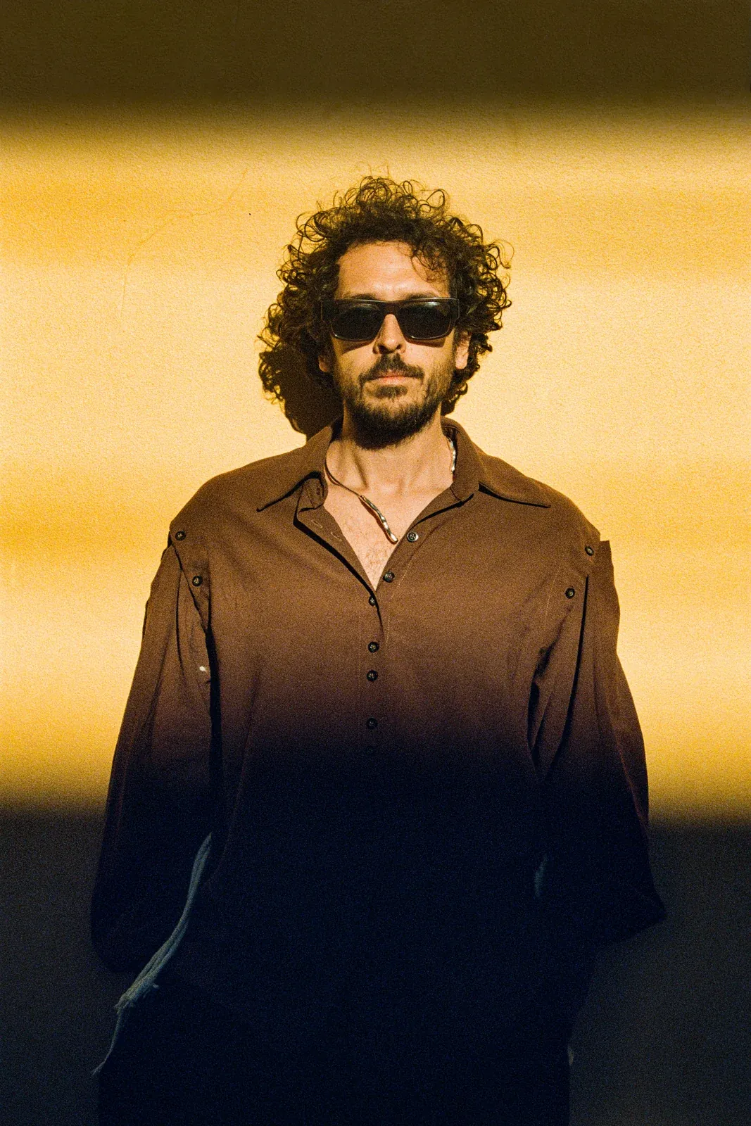 Dobao wearing sunglasses and a brown shirt against a sunlit yellow wall