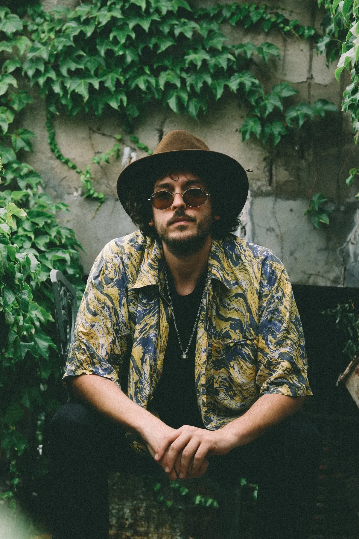 Dobao sitting under ivy, wide hat and round sunglasses