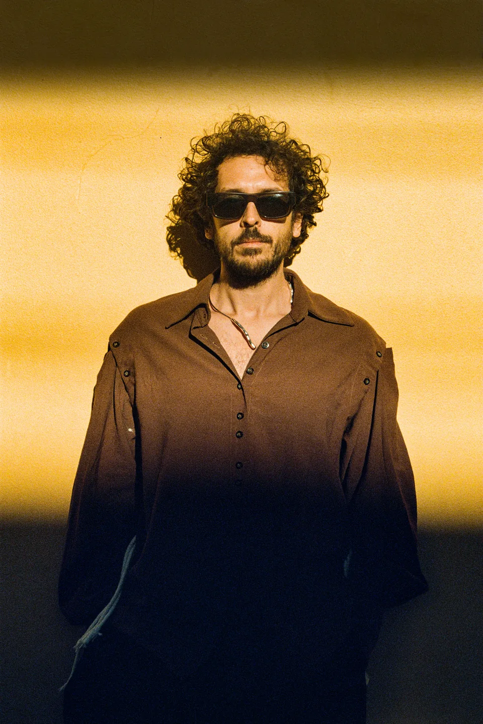 Dobao wearing sunglasses and a brown shirt against a sunlit yellow wall