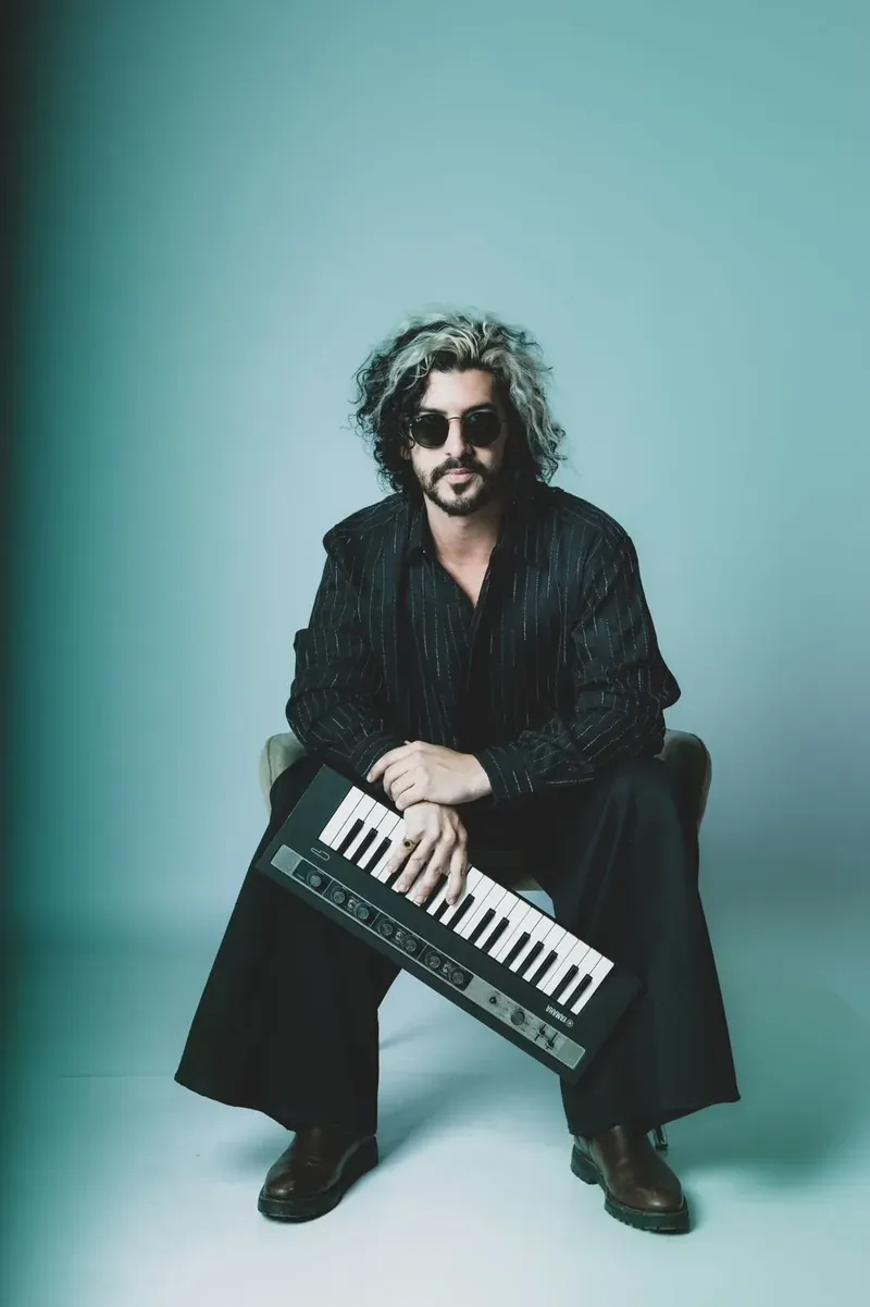 Signo standing with a keytar, black striped shirt, sunglasses